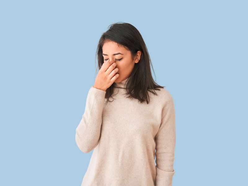 A woman wearing a cream shirt is pinching the bridge of her nose in discomfort