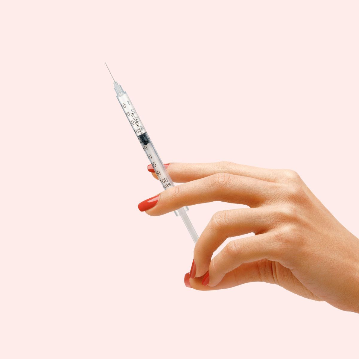 A woman's hand with red nails is holding a syringe in front of a light pink background