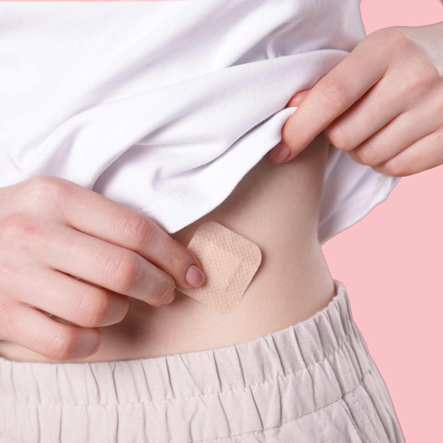 a woman in neutral clothing applying a birth control patch to her stomach against a pink background