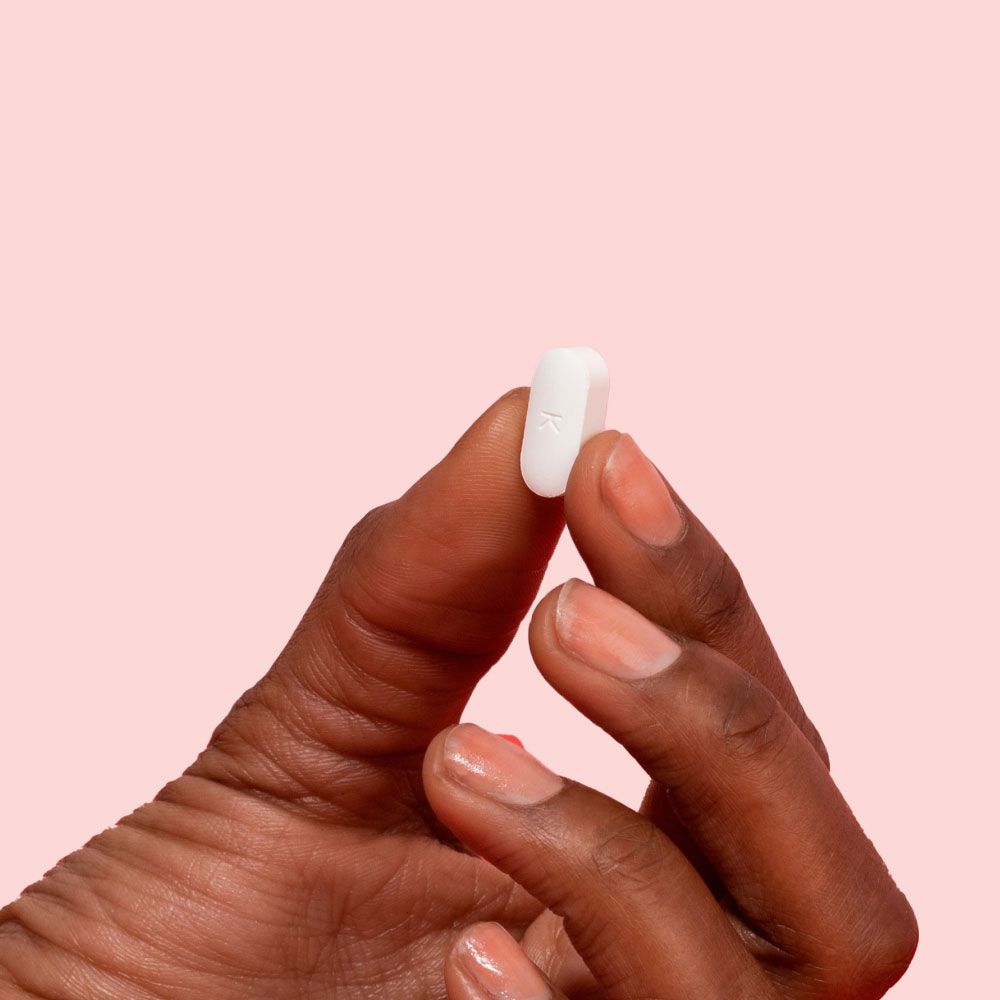 A close up of a woman's hand holding a white pill in front of a pink background