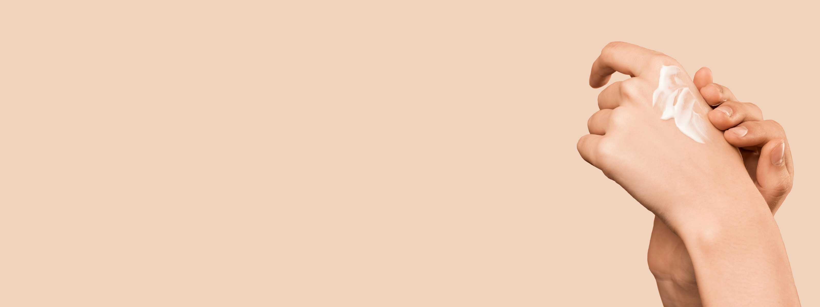 Hands applying white moisturizer or lotion against a peach background.