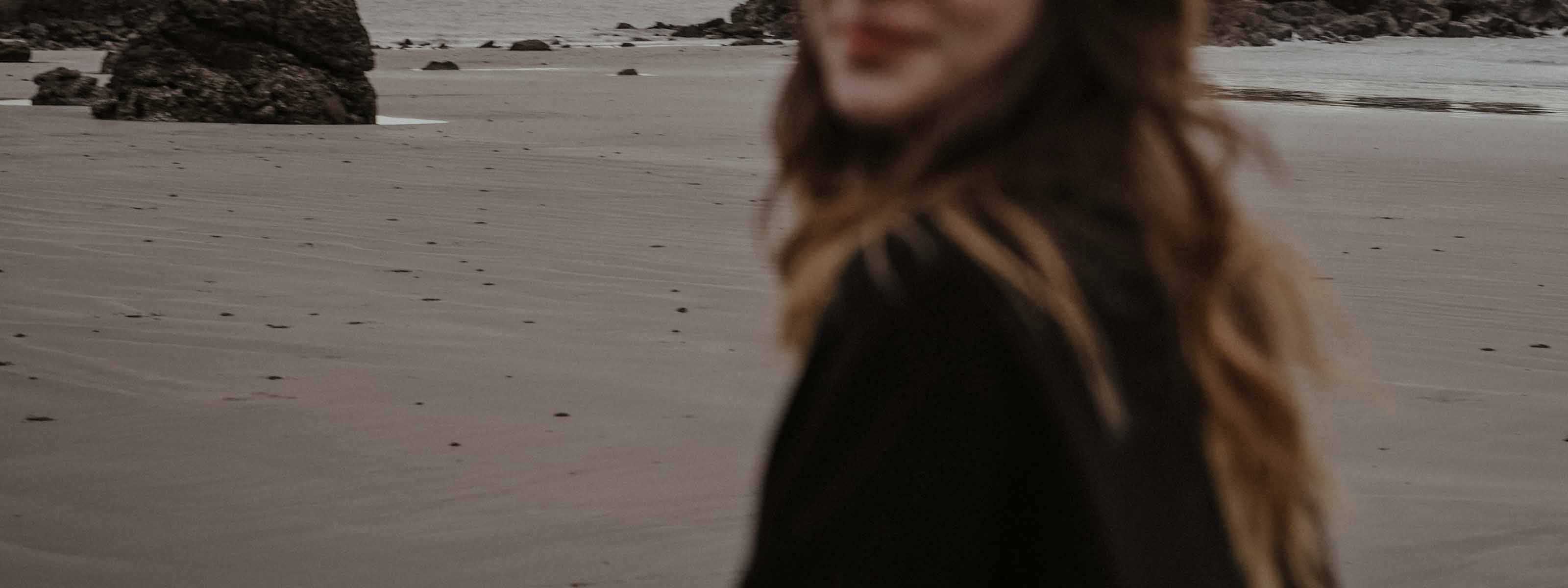 Woman with windswept hair in a dark coat on a grey, overcast beach with rocky sea stacks.