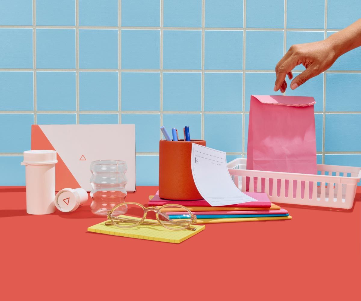 A woman's hand reaching for a pink shopping bag next to an rx slip, pen jar, glasses, a yellow notepad, Wisp medication bottles and diagnostics box on a red surface