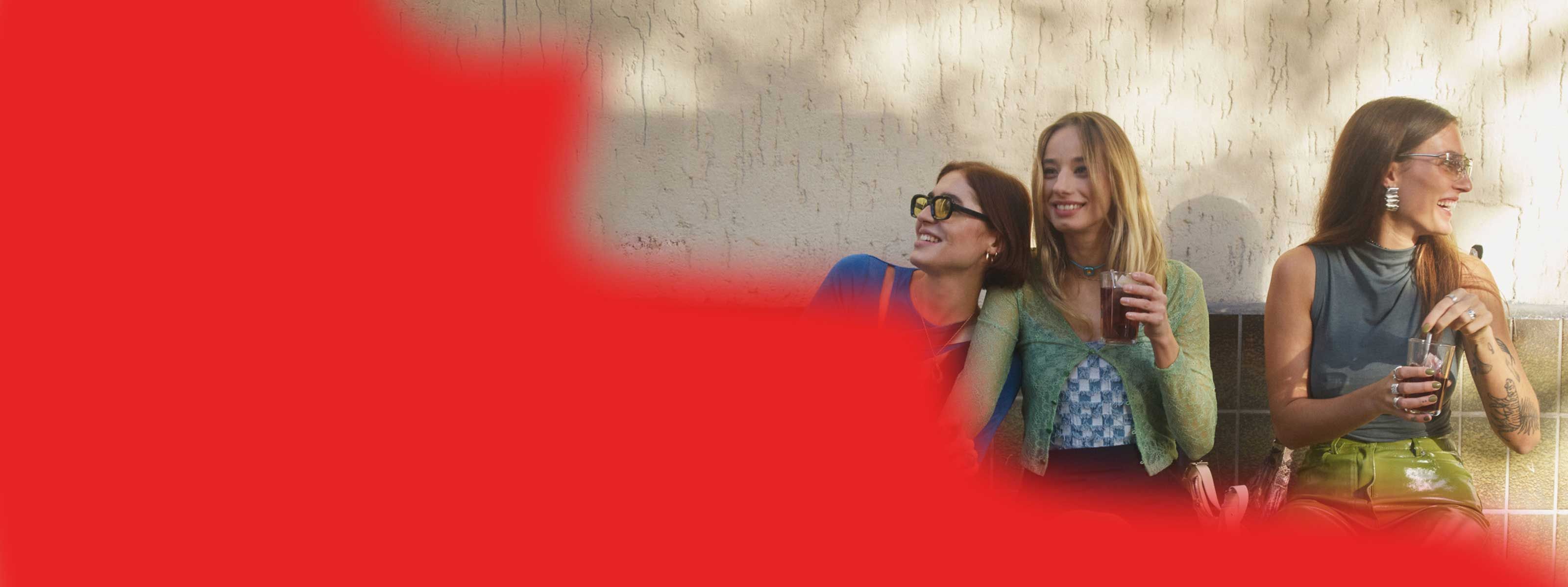 Three fashionable women laughing on a bench with their drinks looking happy