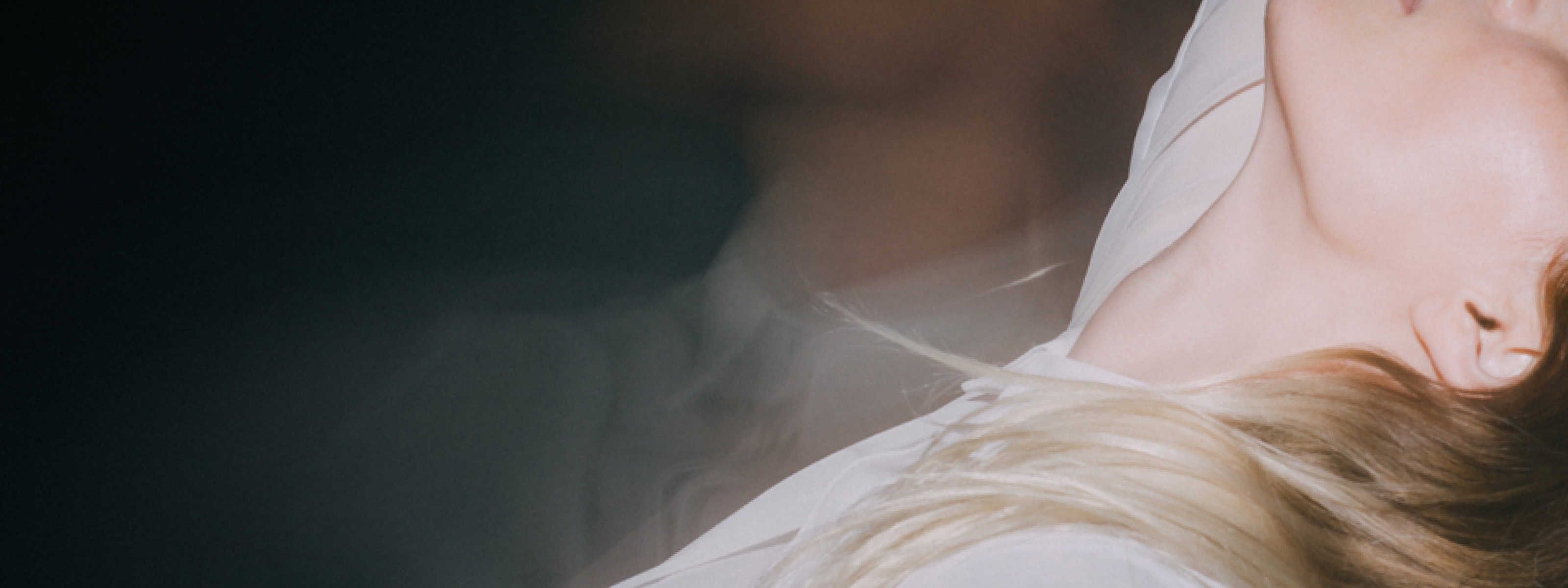 image of a woman's left side profile with a cast of light on her cheek, neck and blonde hair wearing a white shirt and a blurry image of a mans neck and chest in the background