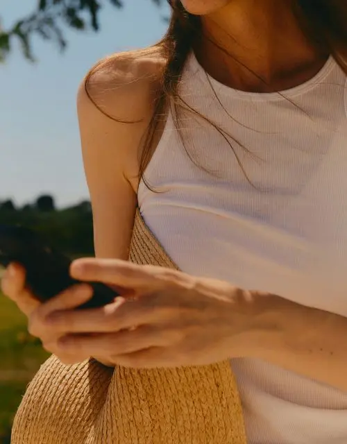 image of a woman in a white shirt and a straw hat under her arm while she holds a phone and is texting