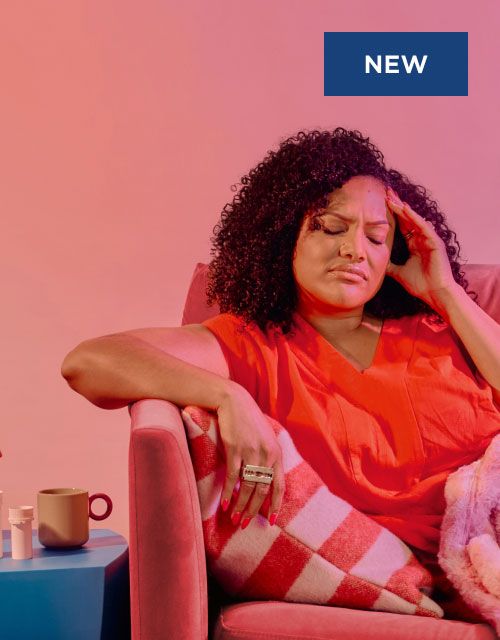 A woman wearing a red shirt it sitting on a pink sofa with her hand on her temple in obvious discomfort