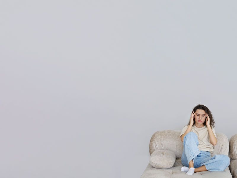 A woman wearing a yellow shirt and blue jeans is sitting on a sofa with her hands on her temples