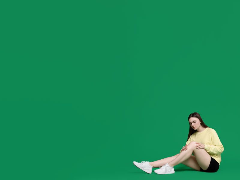 A woman wearing a yellow sweater, black shorts and white sneakers is sitting on a green surface with her hands on her knee in discomfort