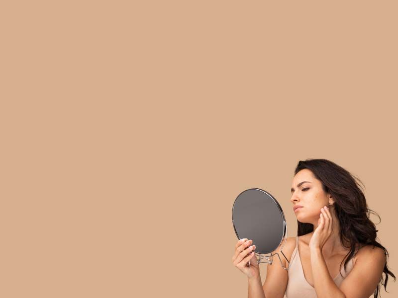 A woman touching her face and looking at her skin in a hand mirror