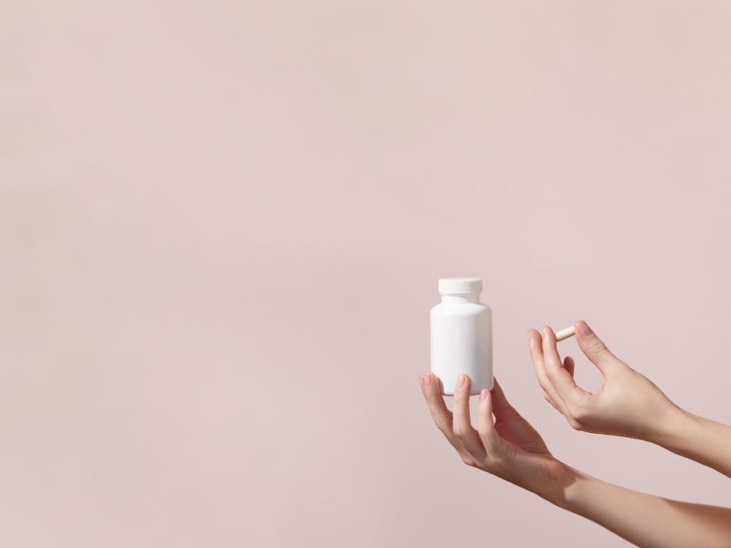 A woman's hands holding a white pill bottle and a supplement capsule in front of a mauve background