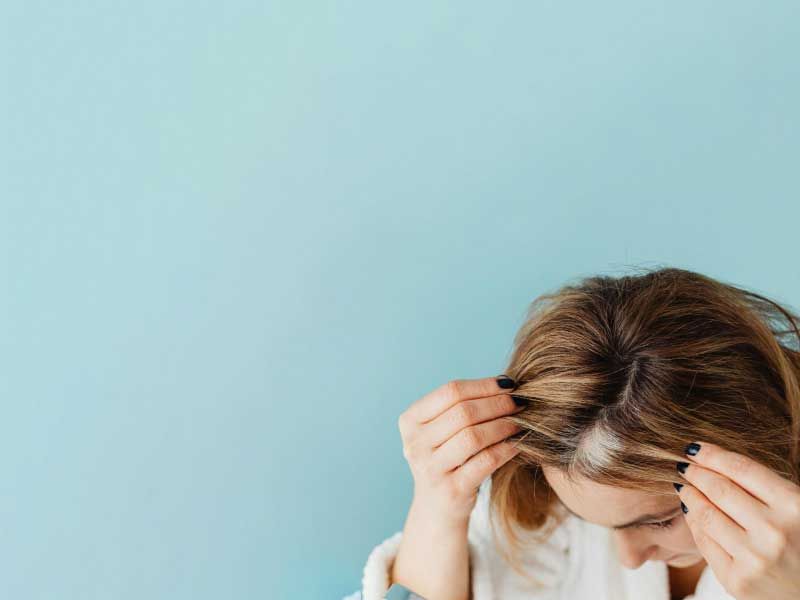 A woman wearing a white robe is looking at her thinning hair in the mirror with a teal background