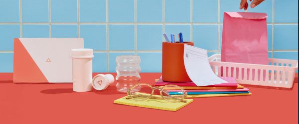 A woman's hand reaching for a pink shopping bag next to an rx slip, pen jar, glasses, a yellow notepad, Wisp medication bottles and diagnostics box on a red surface