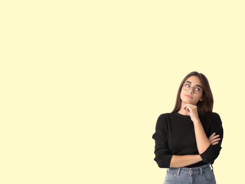 A woman wearing a black sweater and gray jeans is looking pensive in front of a yellow background