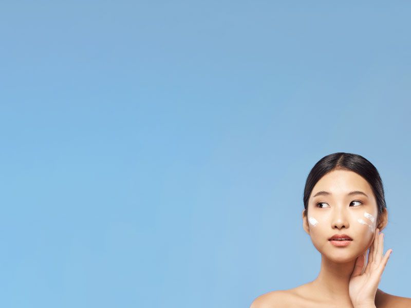 A pretty woman with glowing skin is applying tretinoin cream to her face in front of a blue background