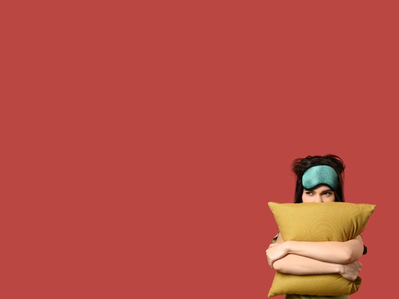 A woman wearing a green sleep mask and hugging a yellow pillow in front of a dark red background