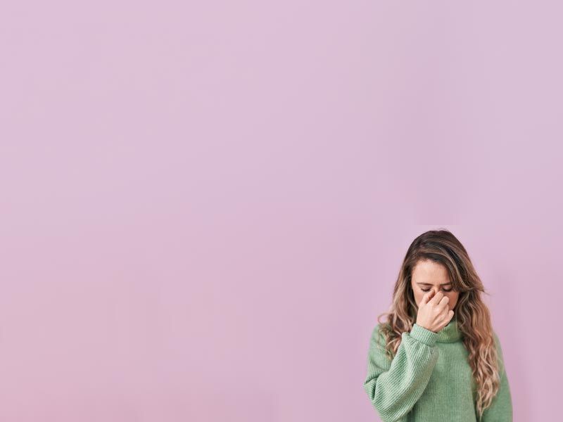 A woman wearing a green sweater has her fingers pinching the bridge of her nose in front of a purple background