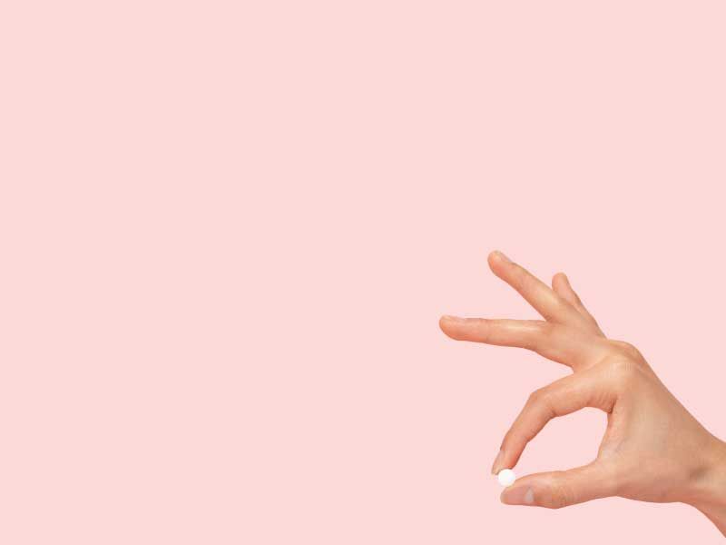A hand holding a small round white pill between the index finger and thumb with a pink background
