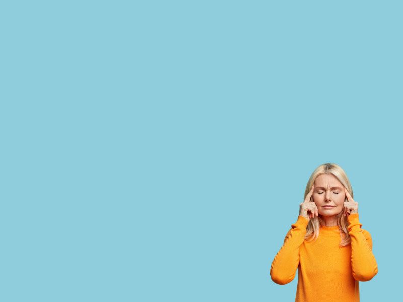 A woman with blonde hair is wearing a yellow sweater with her fingers on her temples in discomfort in front of a blue background