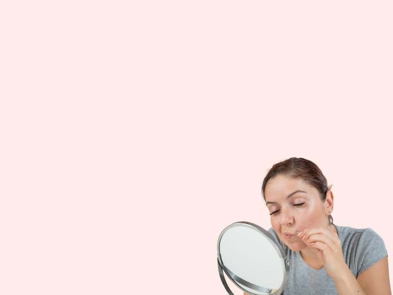 A woman wearing a gray shirt looking at her face in a hand mirror in front of a pink background