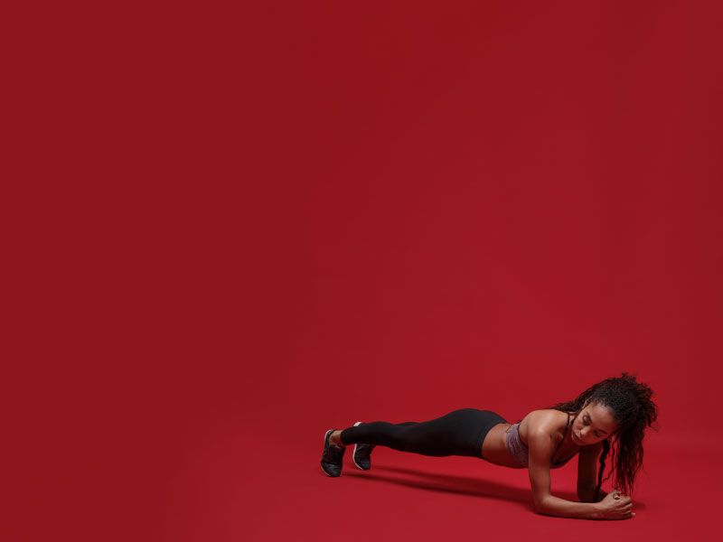 A woman wearing a sports bra, leggings, and sneakers is doing a plank on a red surface