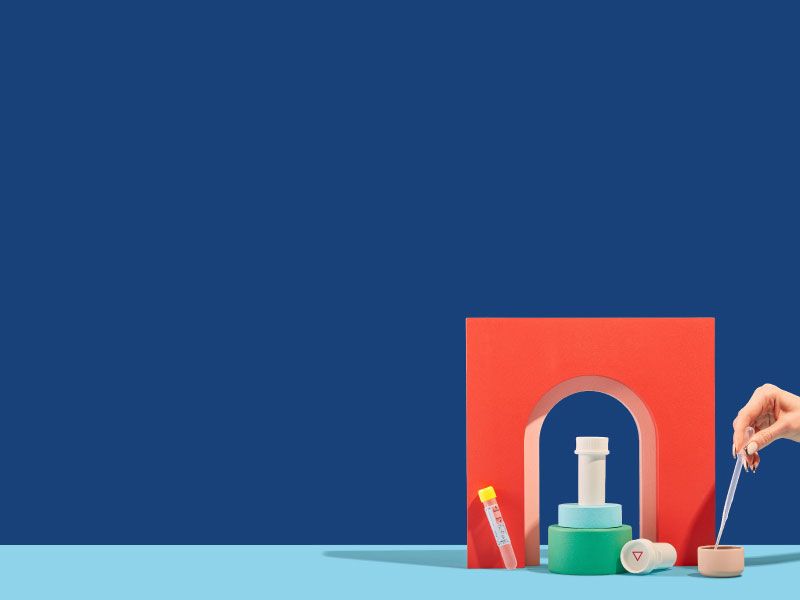 Wisp pill vials, a testing tube, and a woman's hand holding a pipette are balanced on colorful abstract shapes on a light blue surface with a dark blue background