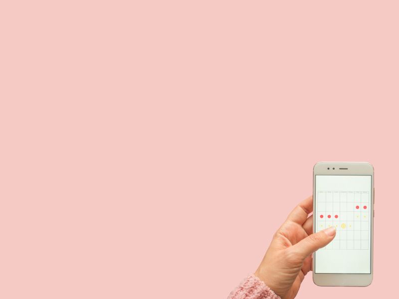 A woman's hand holding a mobile phone with a calendar tracking her menstrual cycle with tampons and menstruation medication on a pink surface in the background