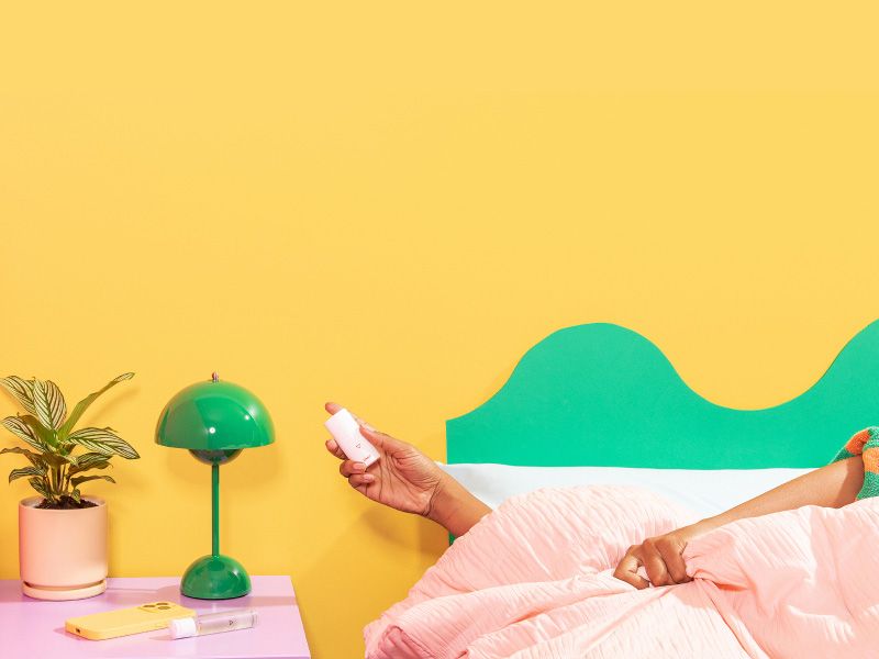 A woman in bed holding Wisp OMG Cream next to a purple nightstand with a green lam, a plant, Wisp Harmonizing Lube, and a mobile phone on it with red and yellow walls