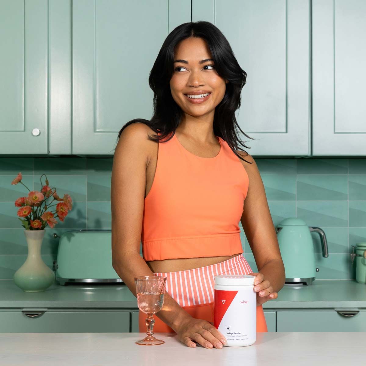 A woman wearing orange fitness clothing is in the kitchen with a bottle of Wisp Revive Collagen Supplements on the counter