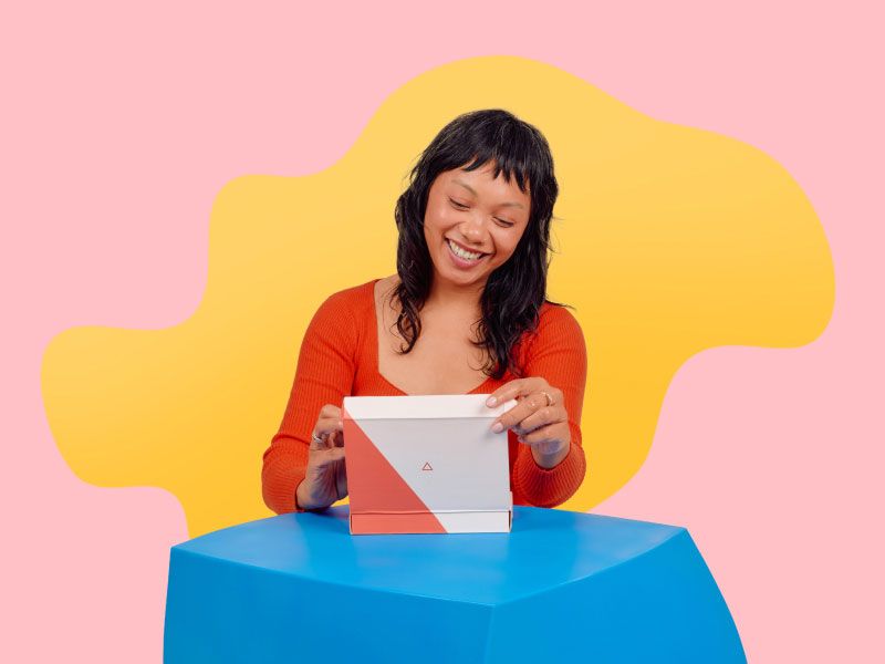 A smiling woman wearing a red top is opening a Wisp Diagnostics package on a blue table