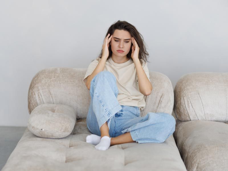 A woman wearing a yellow shirt and blue jeans is sitting on a sofa with her hands on her temples