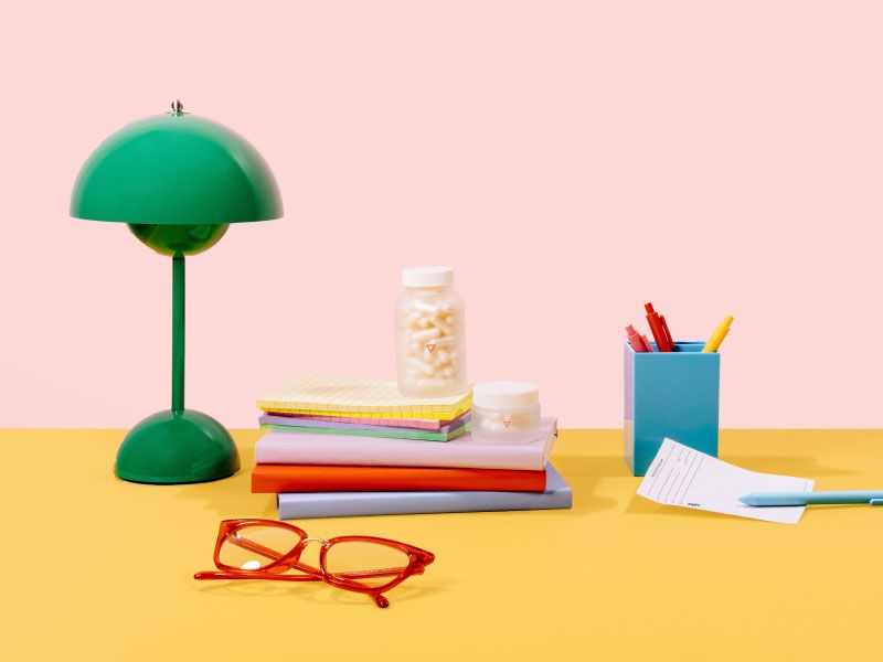 A desktop scene with a green lamp, red glasses, a stack of notebooks and a blue pen holder with an rx script and two Wisp glass jars on a pink and yellow background