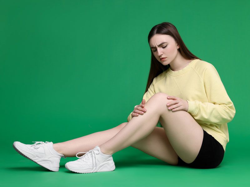 A woman wearing a yellow sweater, black shorts and white sneakers is sitting on a green surface with her hands on her knee in discomfort
