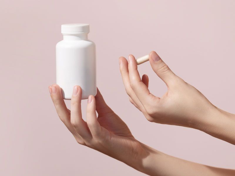 A woman's hands holding a white pill bottle and a supplement capsule in front of a mauve background