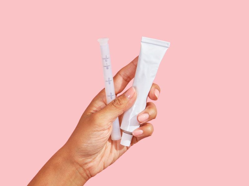 Female hand holding a tube of vaginal dryness cream and applicator with a pink background