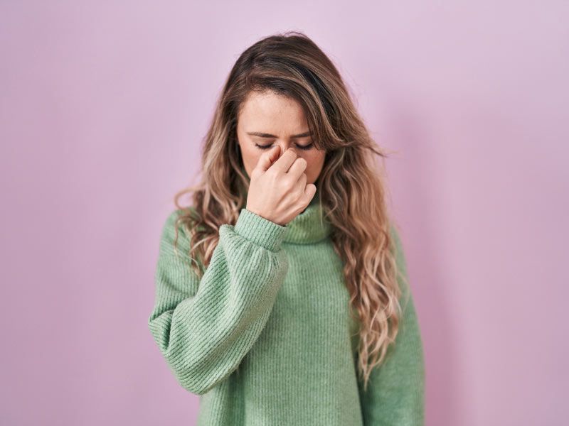 A woman wearing a green sweater has her fingers pinching the bridge of her nose in front of a purple background