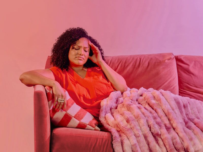 A woman wearing a red shirt is sitting on a fuschia sofa with a pink blanket and she has her hand on her temple in discomfort