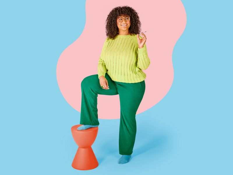 A woman wearing a lime green sweater, dark green pants and light blue socks is smiling and holding a syringe with her foot on on an orange stool