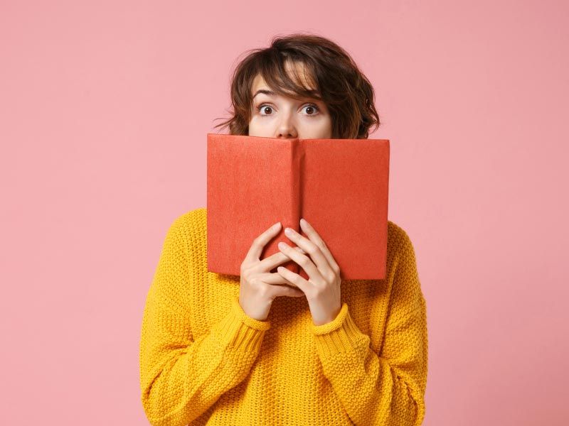 A woman wearing a yellow sweater and holding a brown book with a surprised look on her face