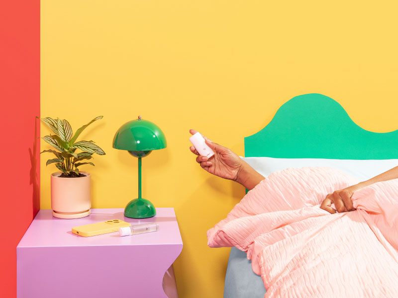 A woman in bed holding Wisp OMG Cream next to a purple nightstand with a green lam, a plant, Wisp Harmonizing Lube, and a mobile phone on it with red and yellow walls