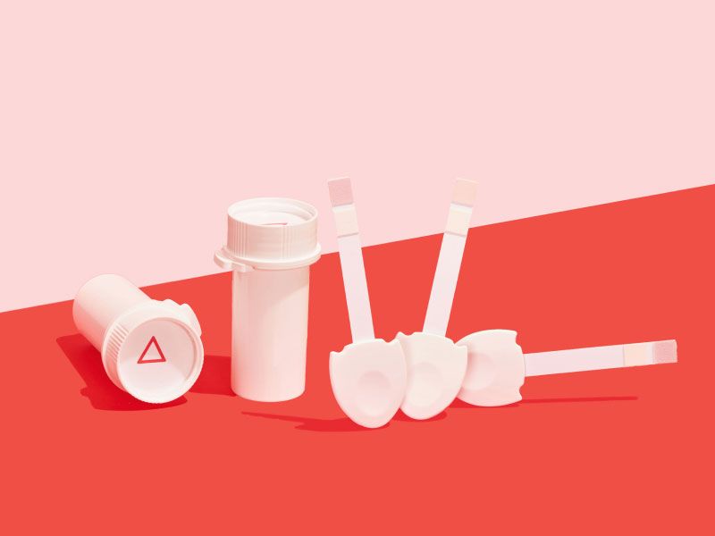 Two Wisp pill vials and 3 urine testing strips on a red surface with a pink background