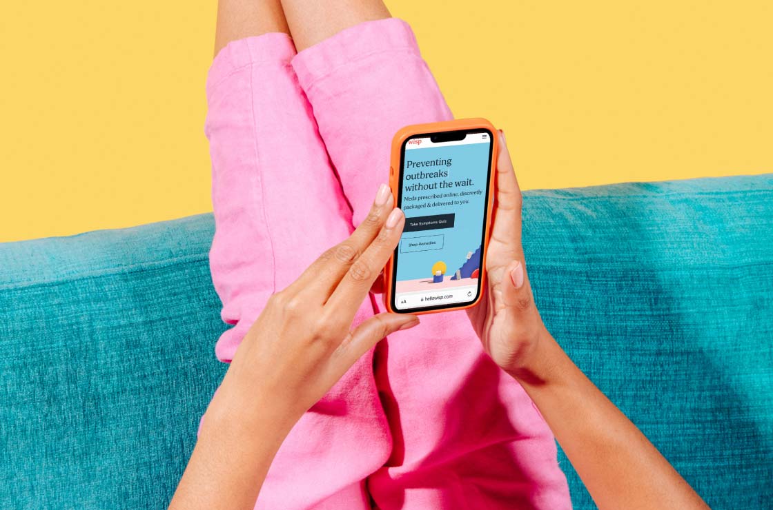 A woman wearing a pink jumpsuit with her feet up on a sofa is holding a phone with Wisp's homepage on it